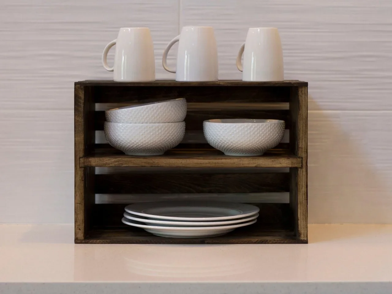 A dark timber crate with a central shelf holds plates and bowls on a pale kitchen counter.