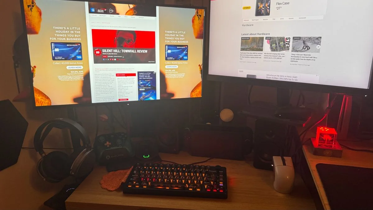 A wood gaming desk with two monitors, a headphone stand, two controllers, a compact lit keyboard and a pale mouse.