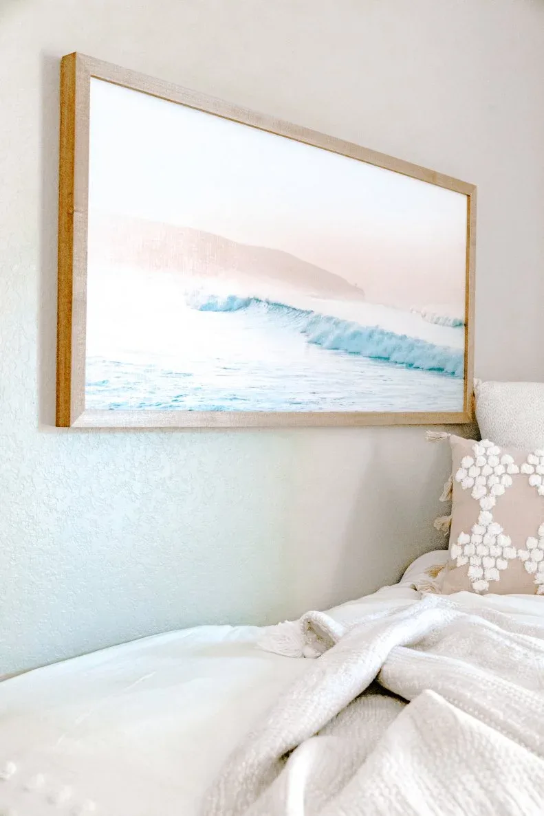Framed pale pink and blue wave picture beside a student bed with white bedding and pale textured pillows.
