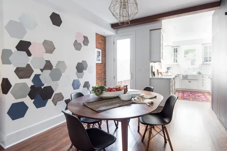 Dining area with a pale wall covered in scattered gray, black, pink, and blue hexagons, oval wooden table, black chairs, and a wire pendant.
