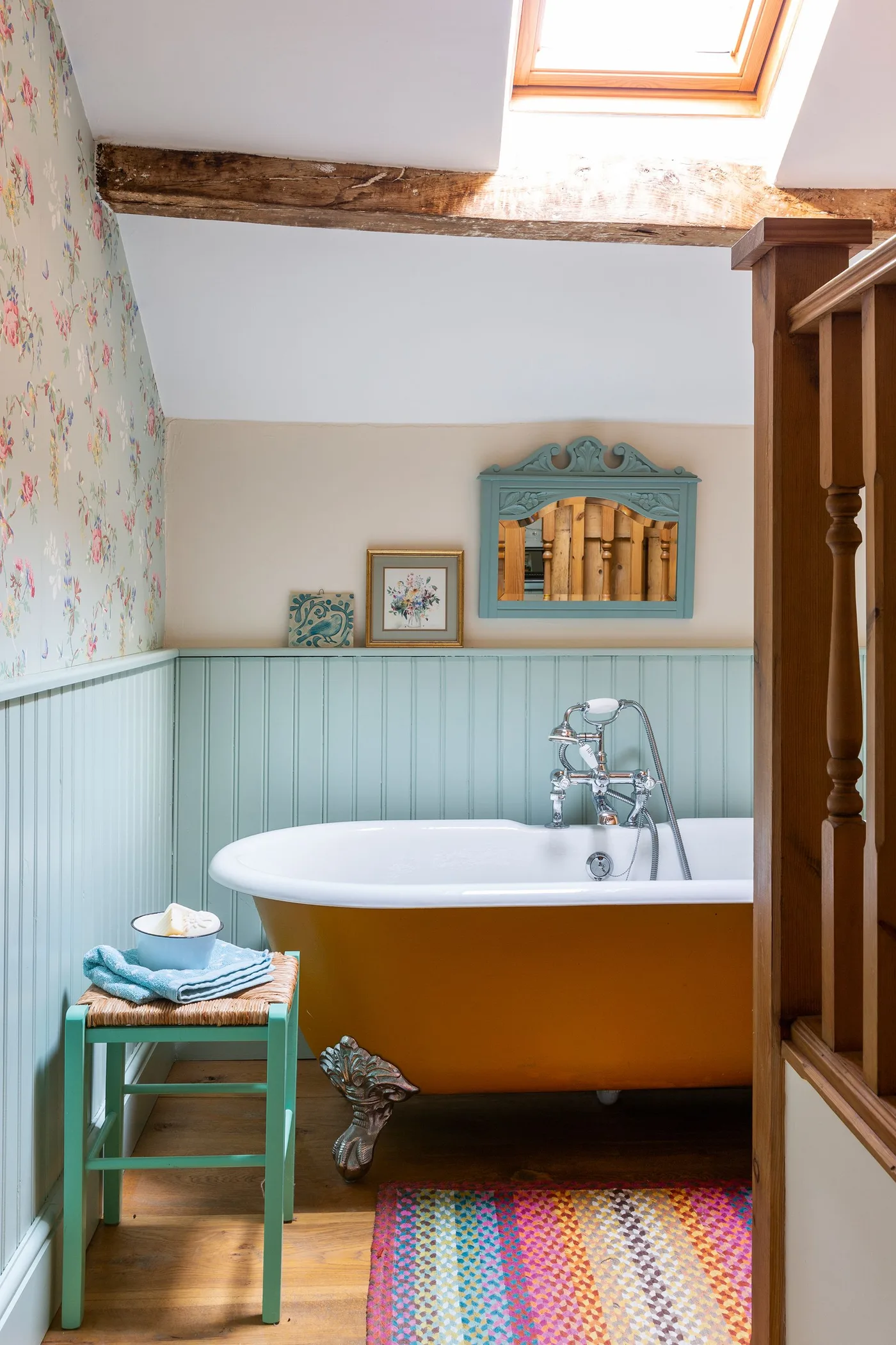 Bathroom with an orange freestanding bath, pale blue paneling, floral upper wall, turquoise mirror and colorful woven rug.