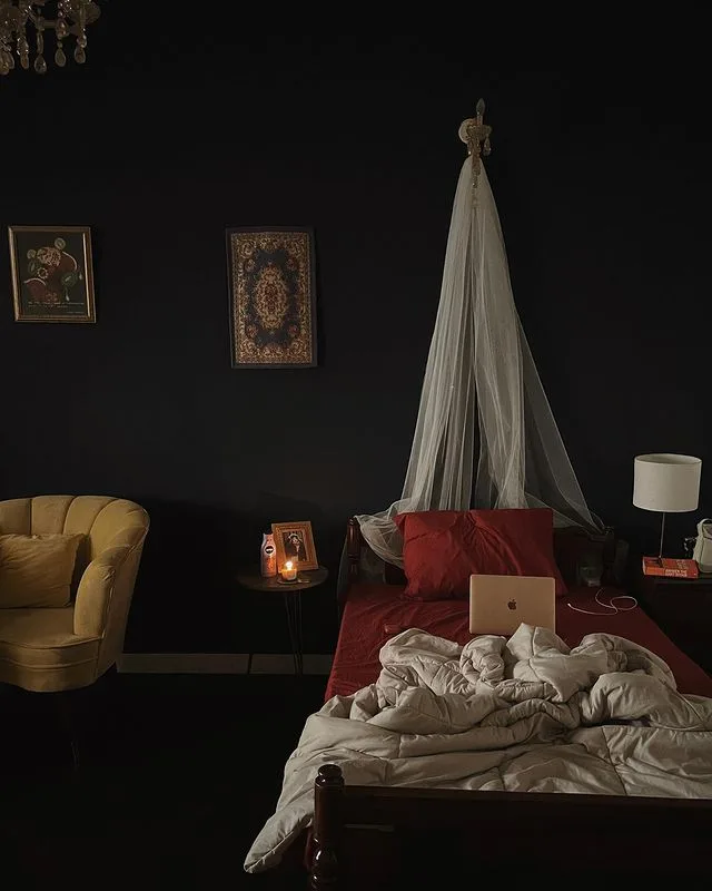 Black bedroom walls with a light hanging canopy, red bedding, framed pictures, yellow chair, and compact bedside tables.