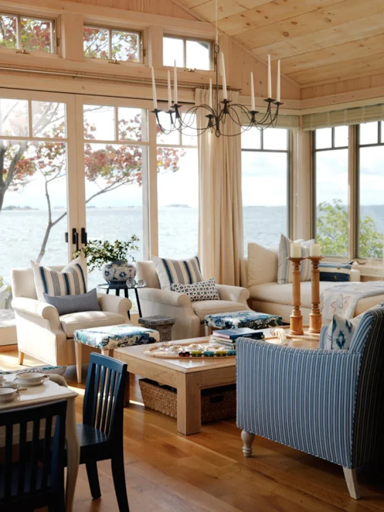 Living room with pale and blue-striped seating, a wooden table, high ceiling and broad waterfront windows.