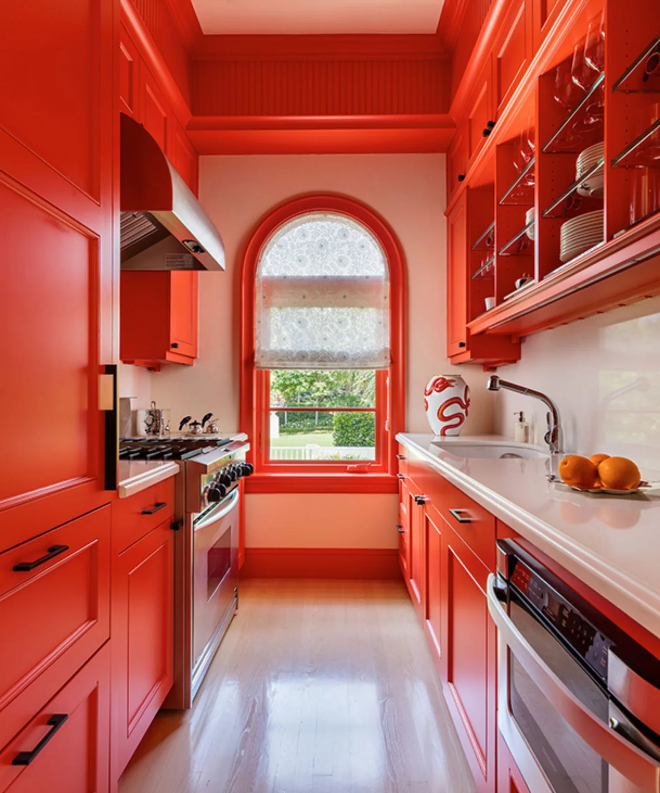 Bright orange cabinets and trim line a narrow kitchen with pale counters, glass-front storage and an arched window with a patterned blind.