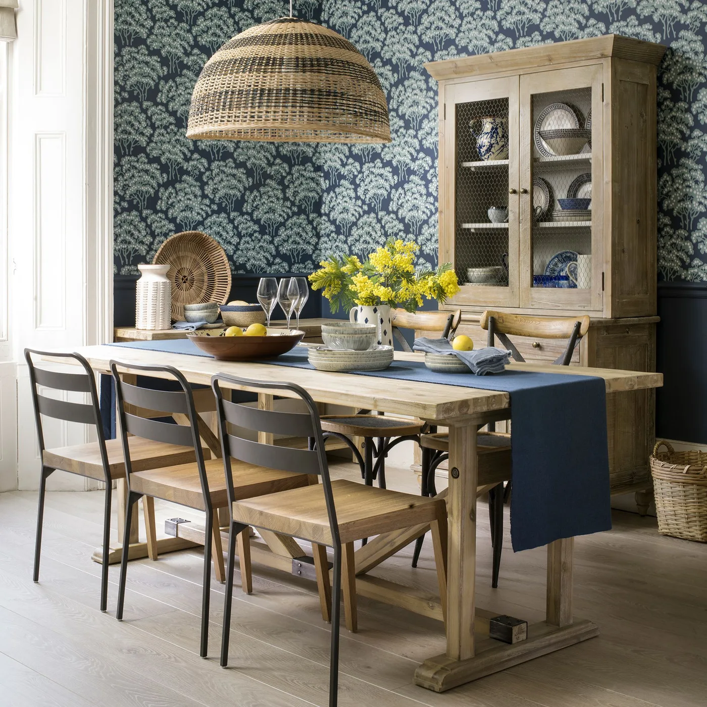 A pale wood table and dish cupboard stand against dark leafy wallpaper below a broad woven pendant.