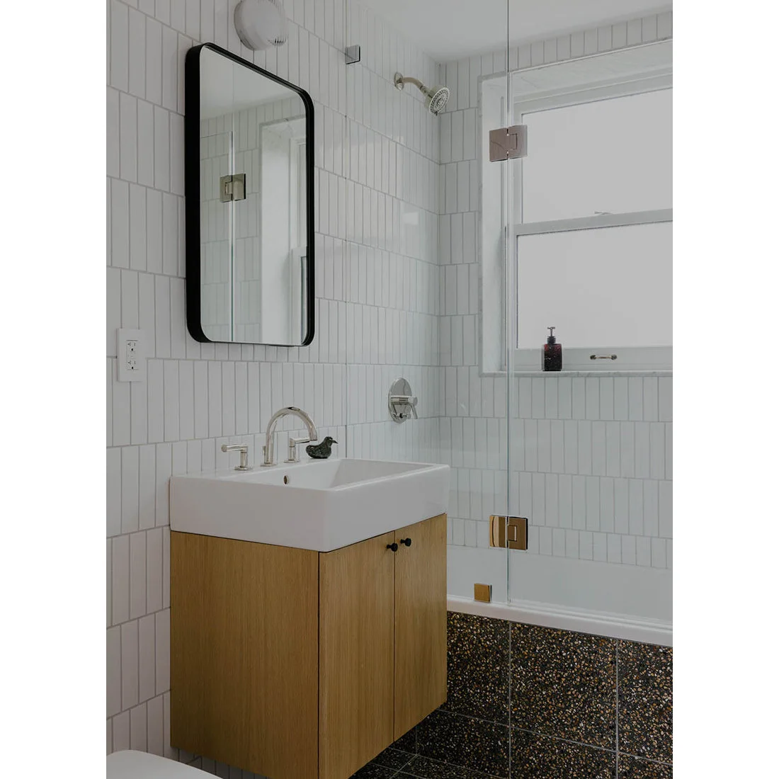 A dark speckled floor and bath front sit beneath white wall tiles beside a compact timber vanity.