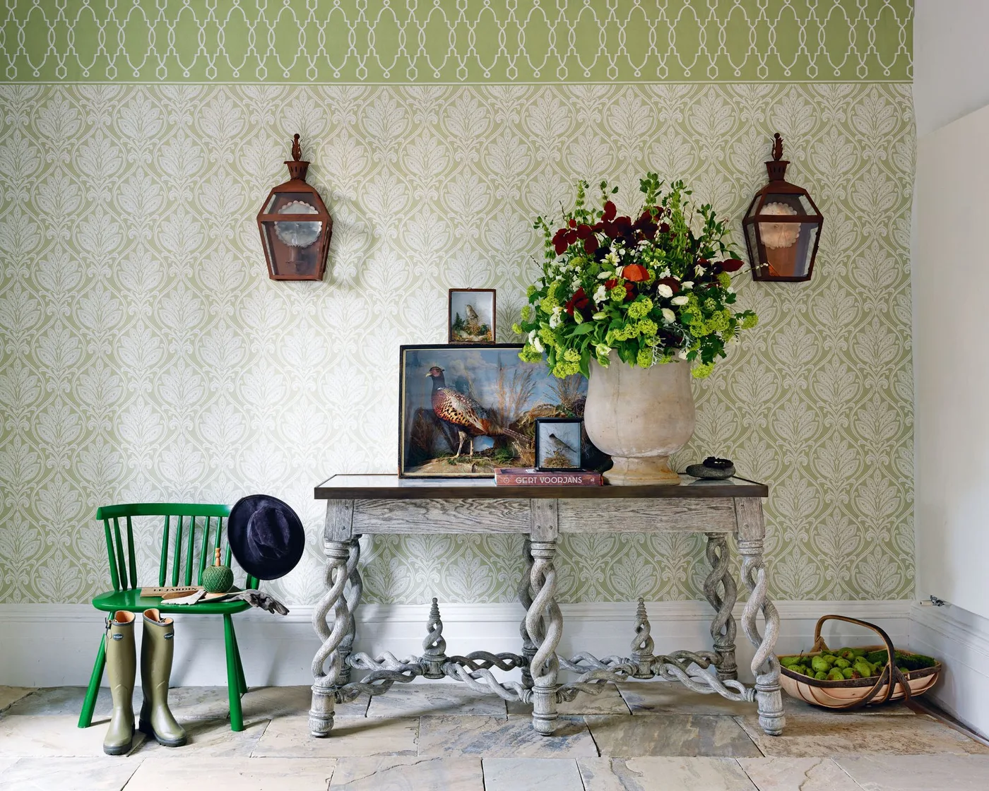 Pale twisted-leg console beneath green patterned wallpaper, with layered bird pictures, flowers, a green chair and wall lanterns