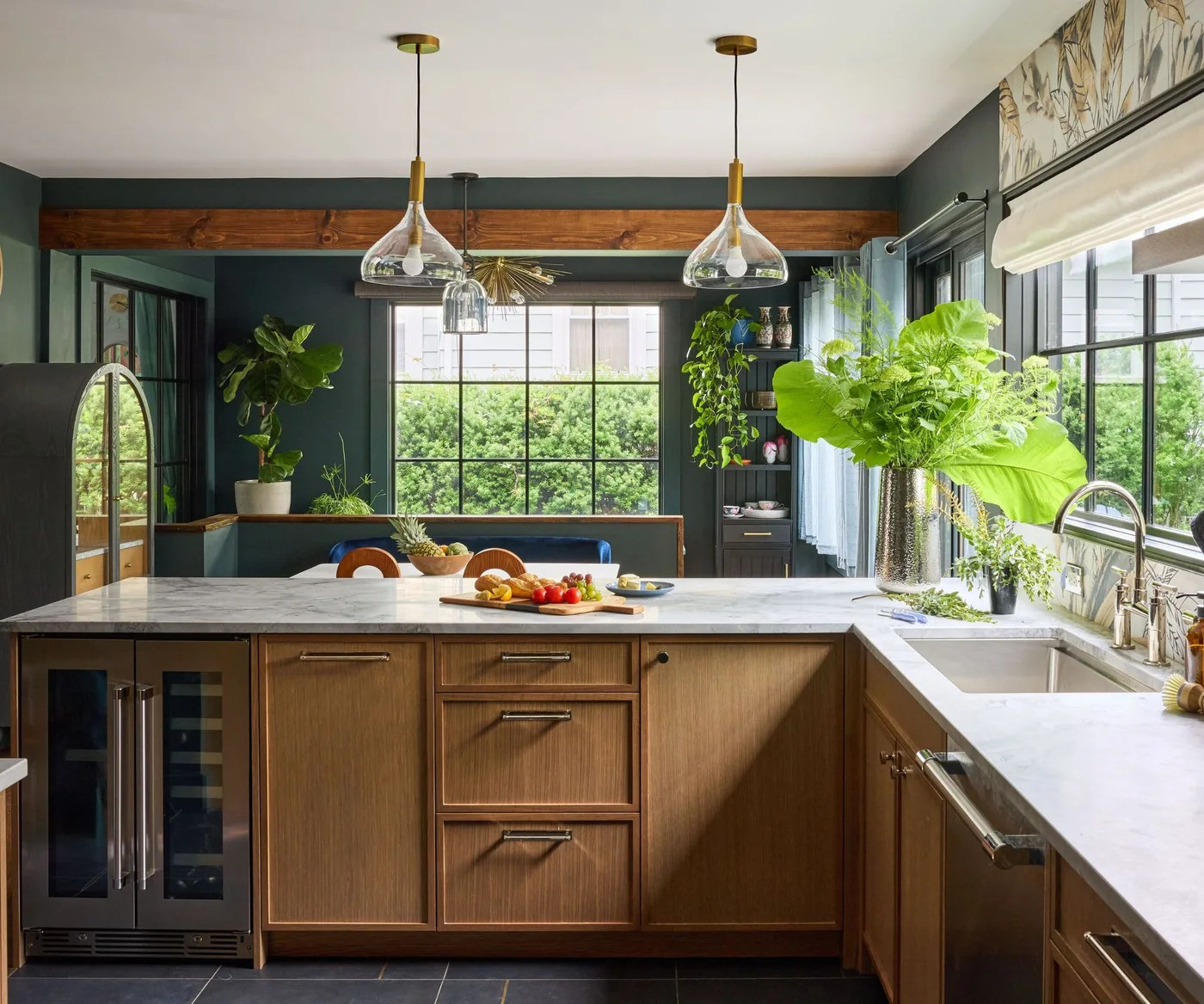 Warm oak peninsula cabinets with a drinks refrigerator face dark walls and a windowed dining nook, with pale counters and glass pendants.