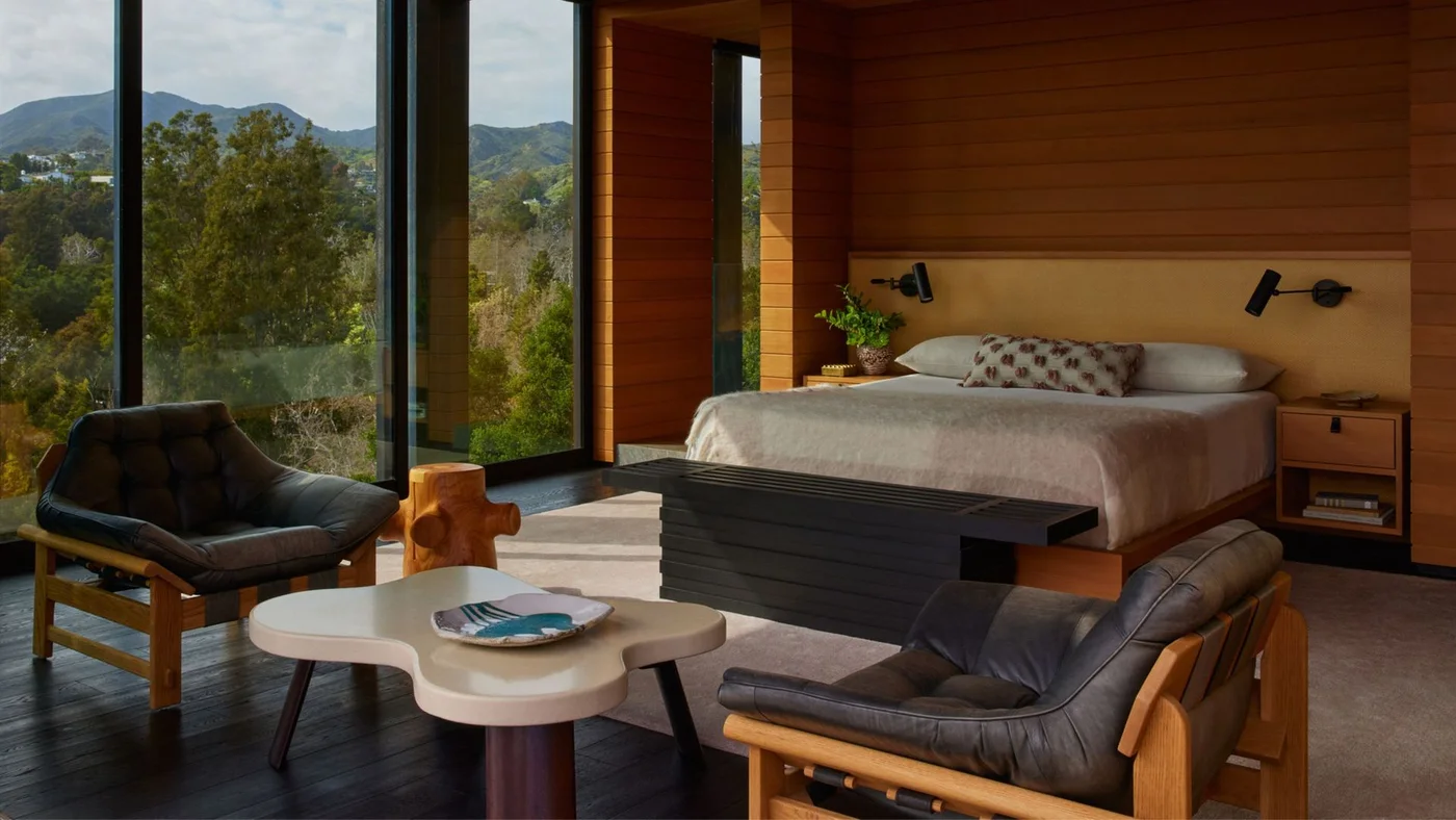Two dark cushioned wood-framed chairs face a pale curved table beside a low bed, warm wood walls and tall bedroom windows