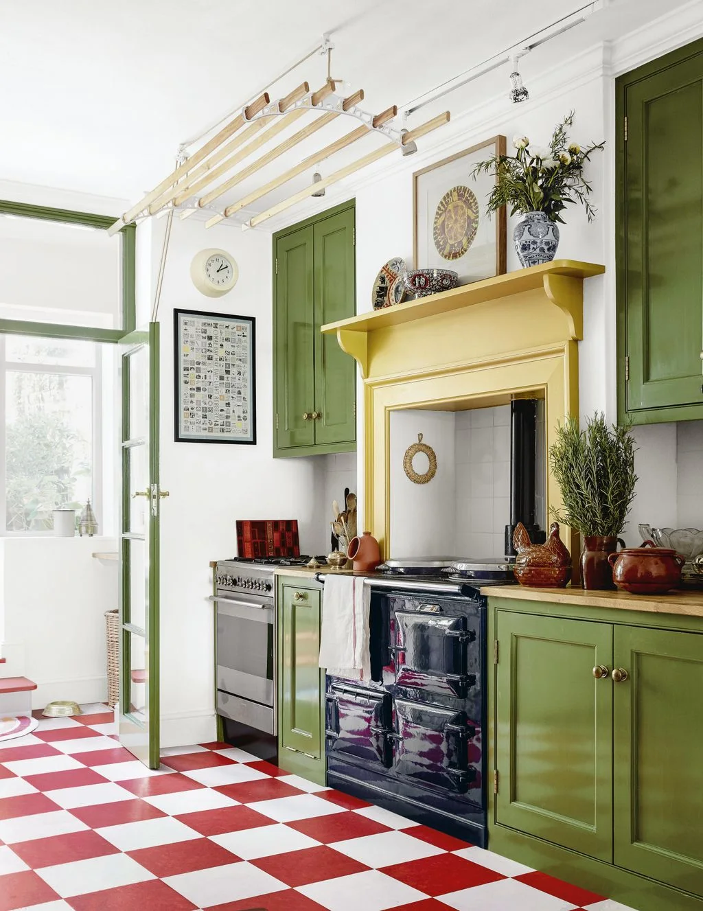Green cabinets and a yellow cooking surround sit above a red-and-white checked floor beside a dark cooker, framed pictures and decorative dishes.