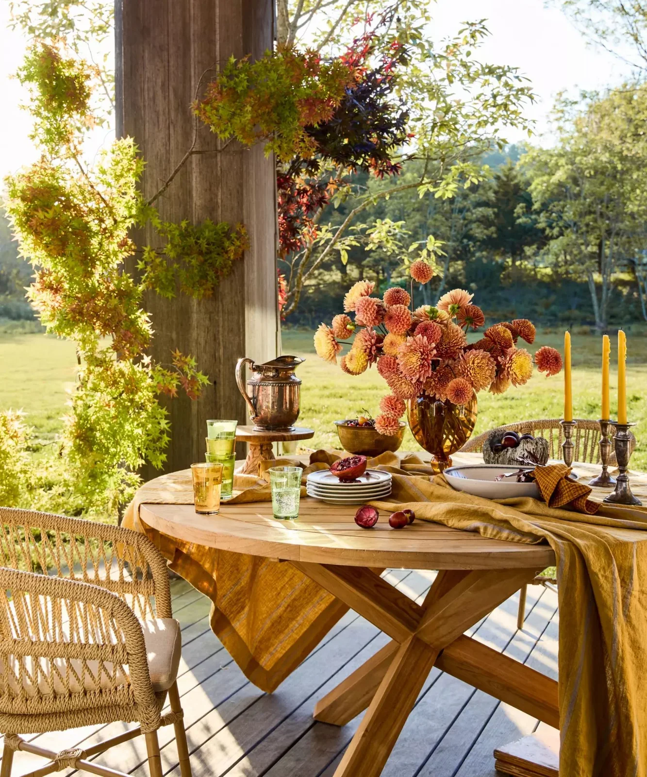 Green and warm-colored foliage follows a porch column beside a round wooden table set with flowers, glasses and pale dishes