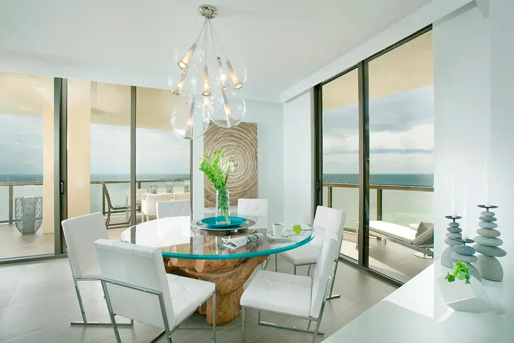 Round clear glass dining table over a sculptural wood-toned base with white chairs beside tall windows