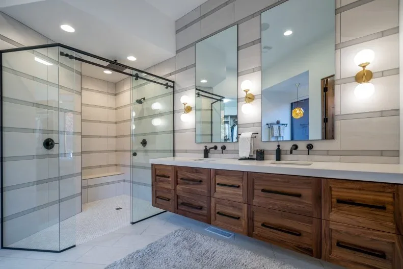 Horizontal pale and gray wall tiles surround a dark-framed shower beside a wood double vanity, large mirrors and gold-colored wall lights