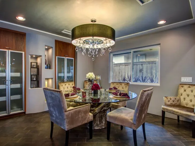 Glass dining table with pale and gray upholstered chairs beneath a drum light with hanging decorative drops.