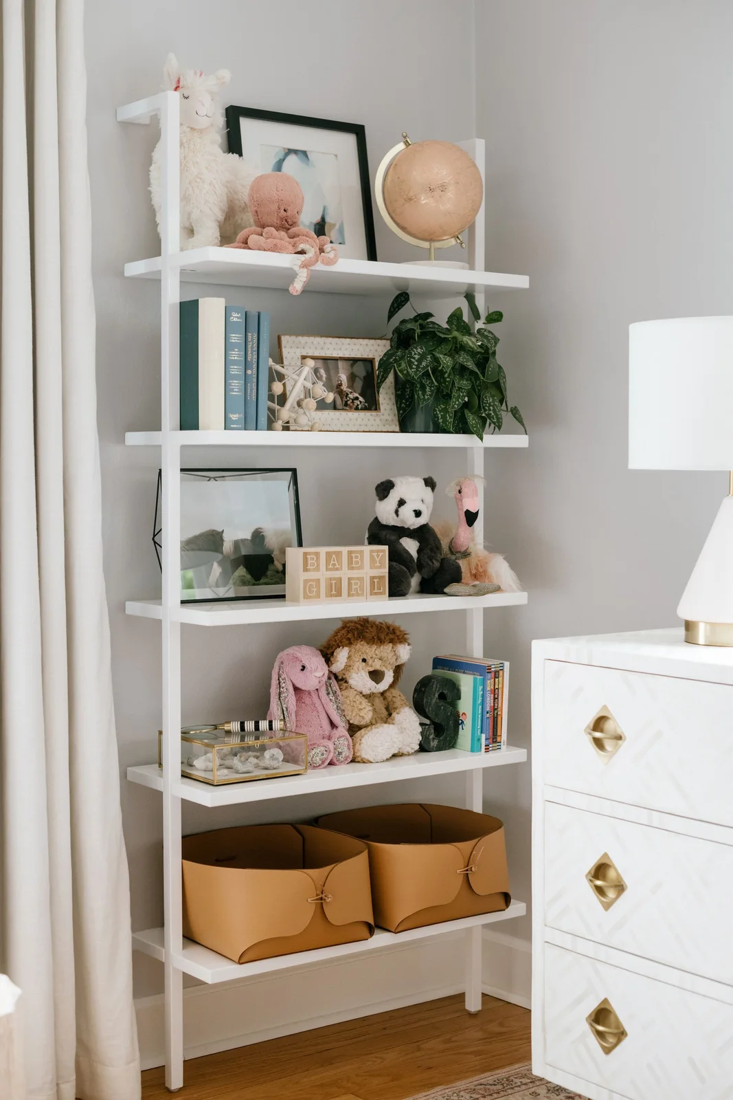 White ladder-style shelving with stuffed animals, books, framed pictures and two tan baskets on the lowest shelf