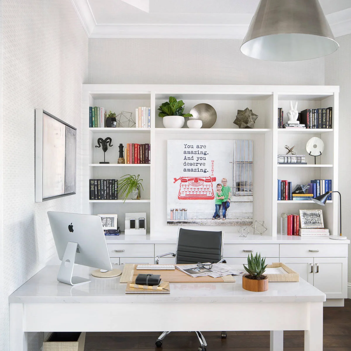 Pale desk with a wood-edged mat, monitor and black chair before white bookshelves