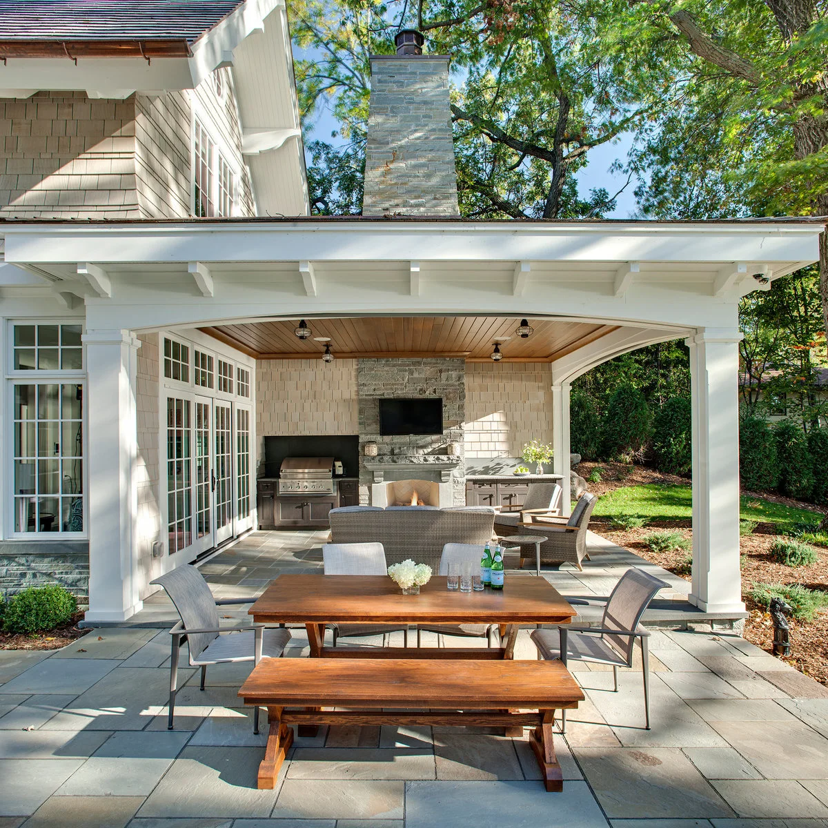 A low white patio roof shelters lounge furniture, while a wood dining table and bench sit on the open paving in front.