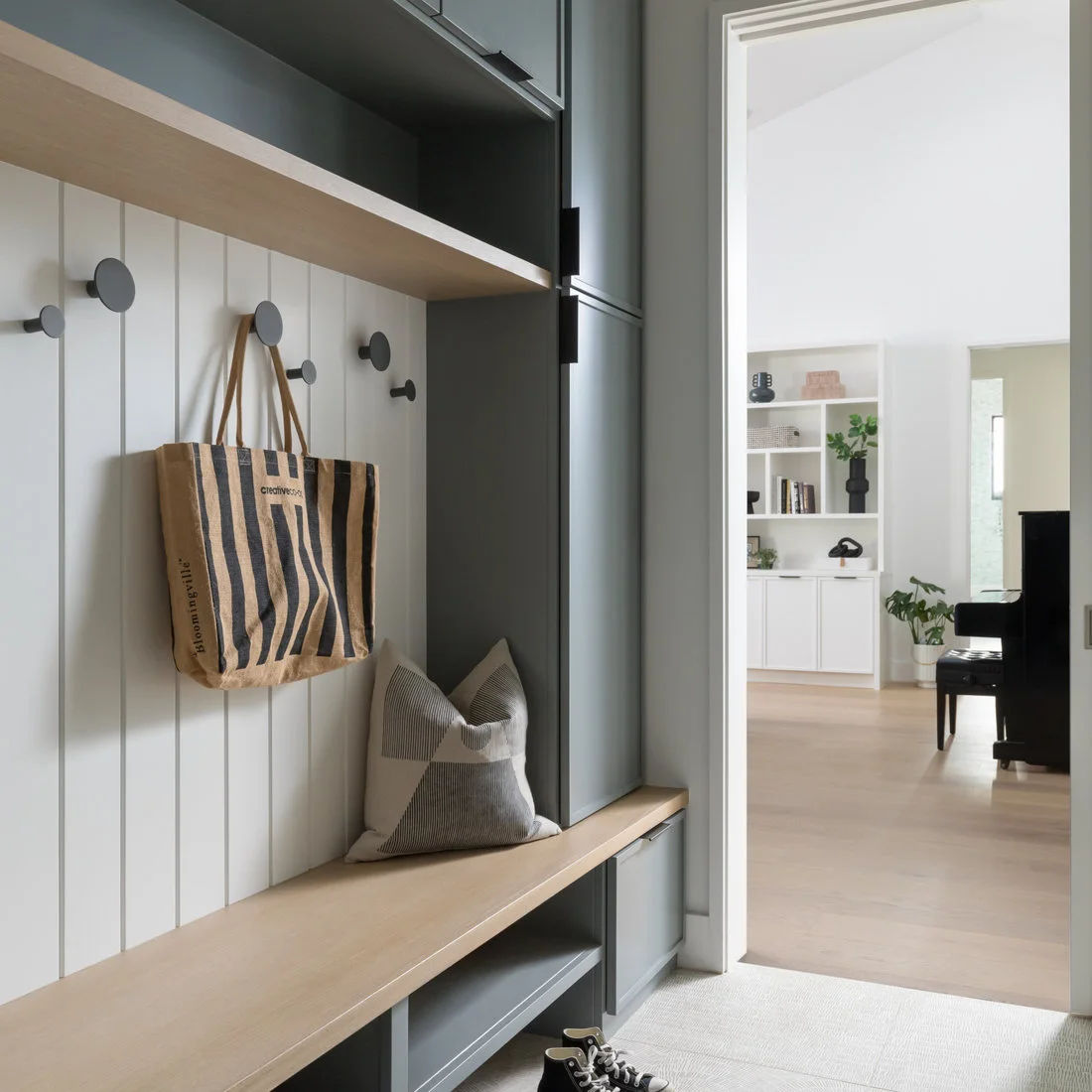 Dark gray entry cupboards frame a pale wood bench and white vertical boards with round gray hooks beside a bright living room.