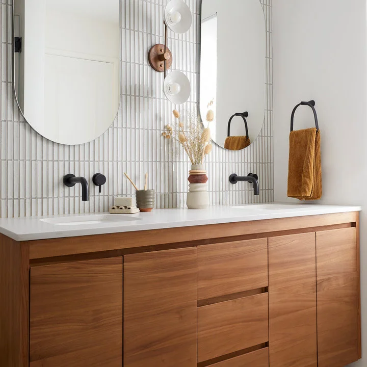 Two mirrors and black wall taps sit above a wood double vanity against narrow vertical pale tiles with mustard towels.