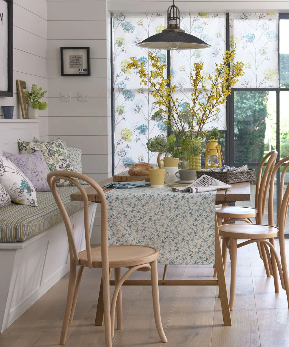 A wood dining table stands beside a cushioned window bench, curved wood chairs, floral blinds and pale paneled walls.