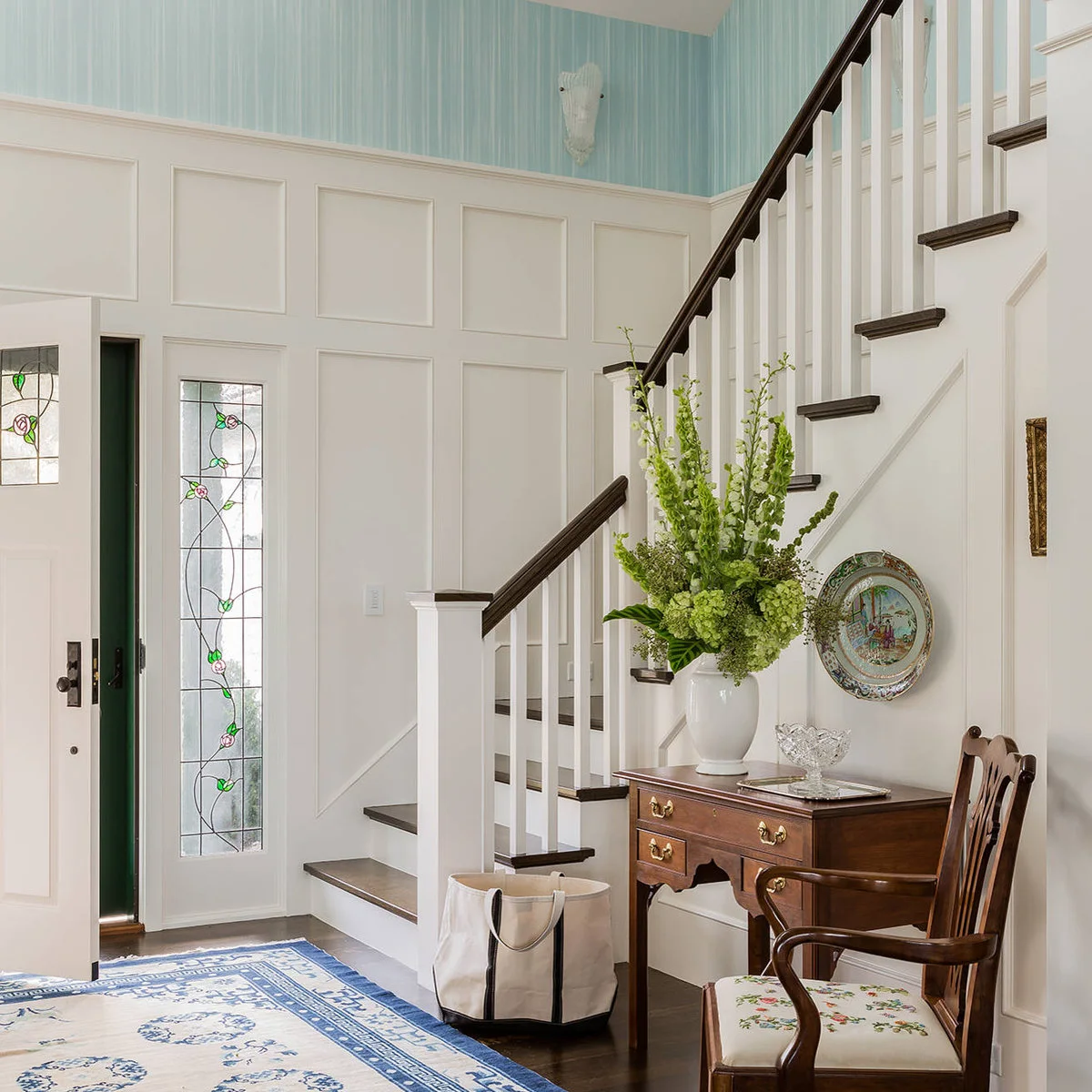 Pale blue striped wallpaper sits above white entry paneling beside a dark-railed staircase.