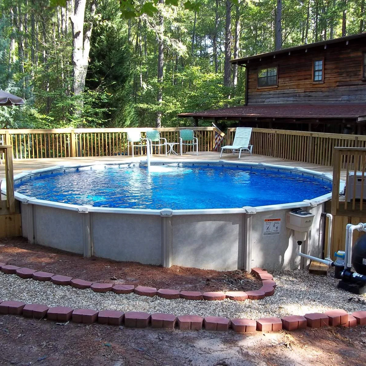 Round gray above-ground pool with a partial wood deck, exposed filter equipment and a curved gravel border.