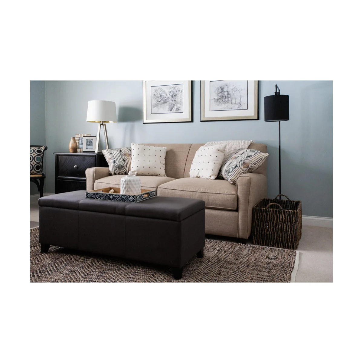 Beige sofa bed with patterned cushions, a dark storage ottoman and a basket beside the arm