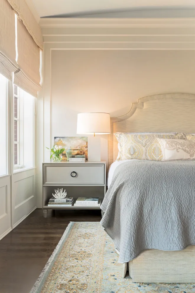 A pale shaped headboard sits beside a gray bedside cabinet under light ceiling beams near a window