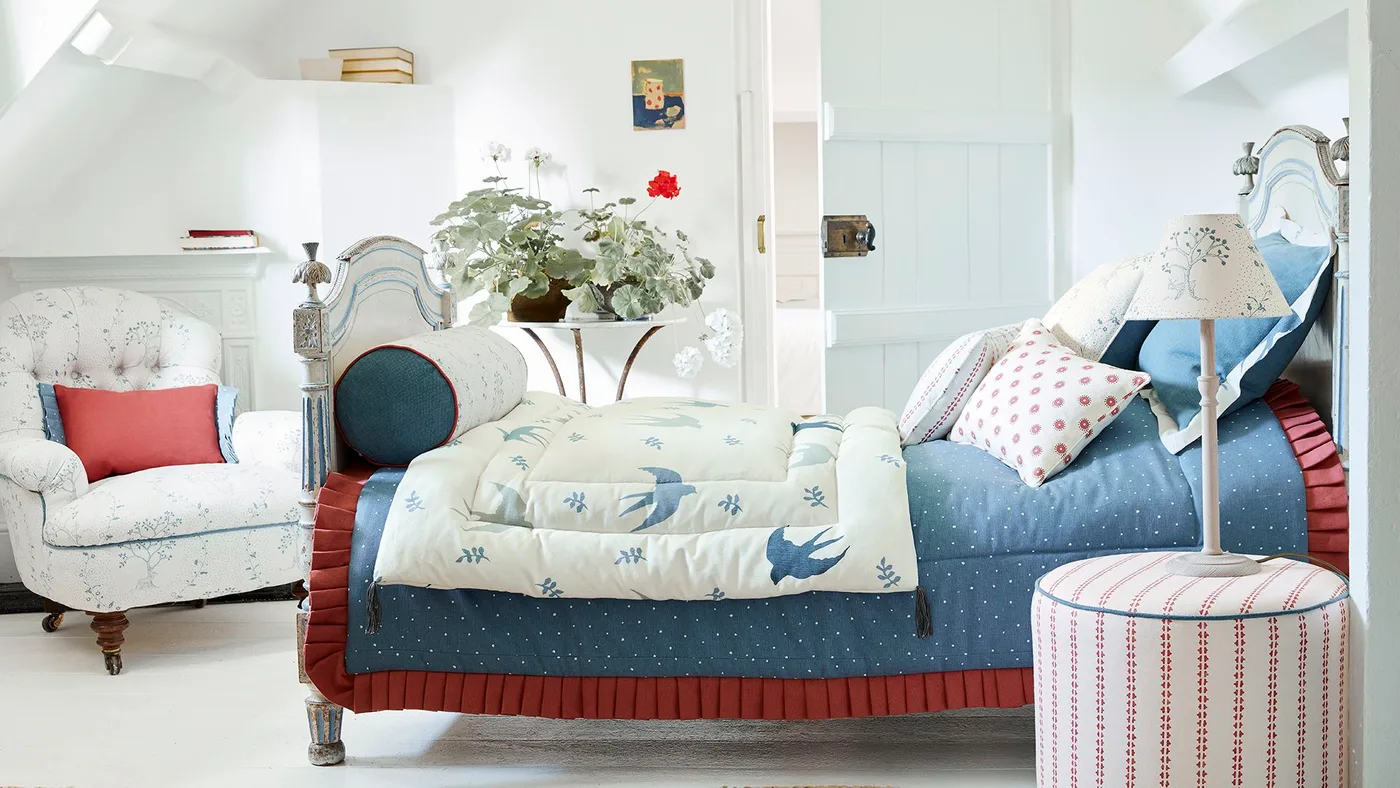 Bright bedroom with a decorative pale bed frame, blue bedding, red bed skirt, patterned armchair and striped round stool.