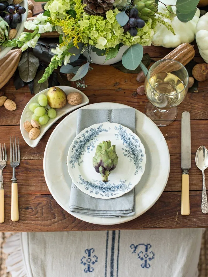 A green artichoke sits on a small patterned plate above a pale napkin beside grapes, flowers and clear glassware