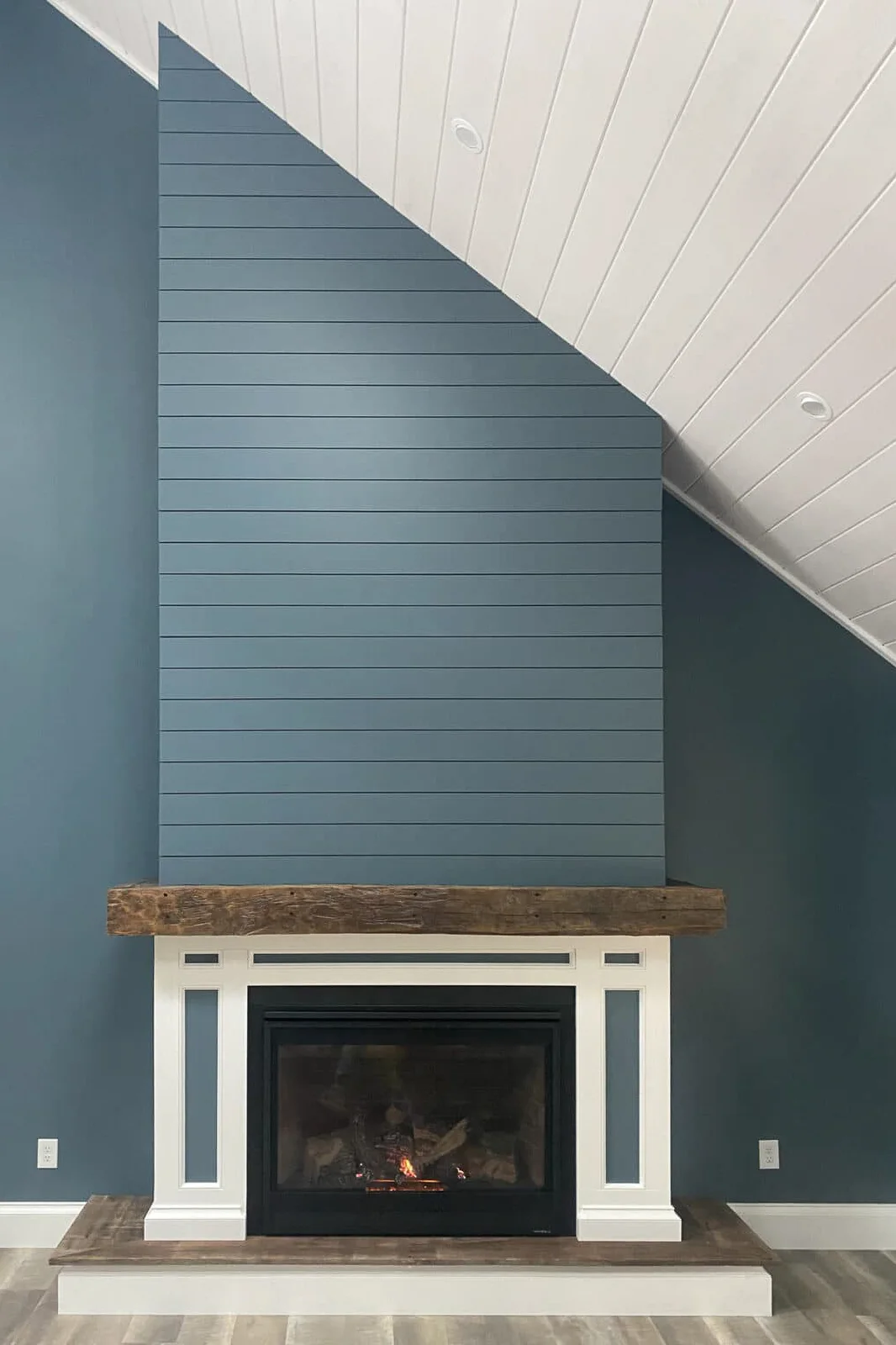 A blue shiplap chimney breast meets a sloping white boarded ceiling above a wood mantel, white trimmed surround and dark fireplace insert.