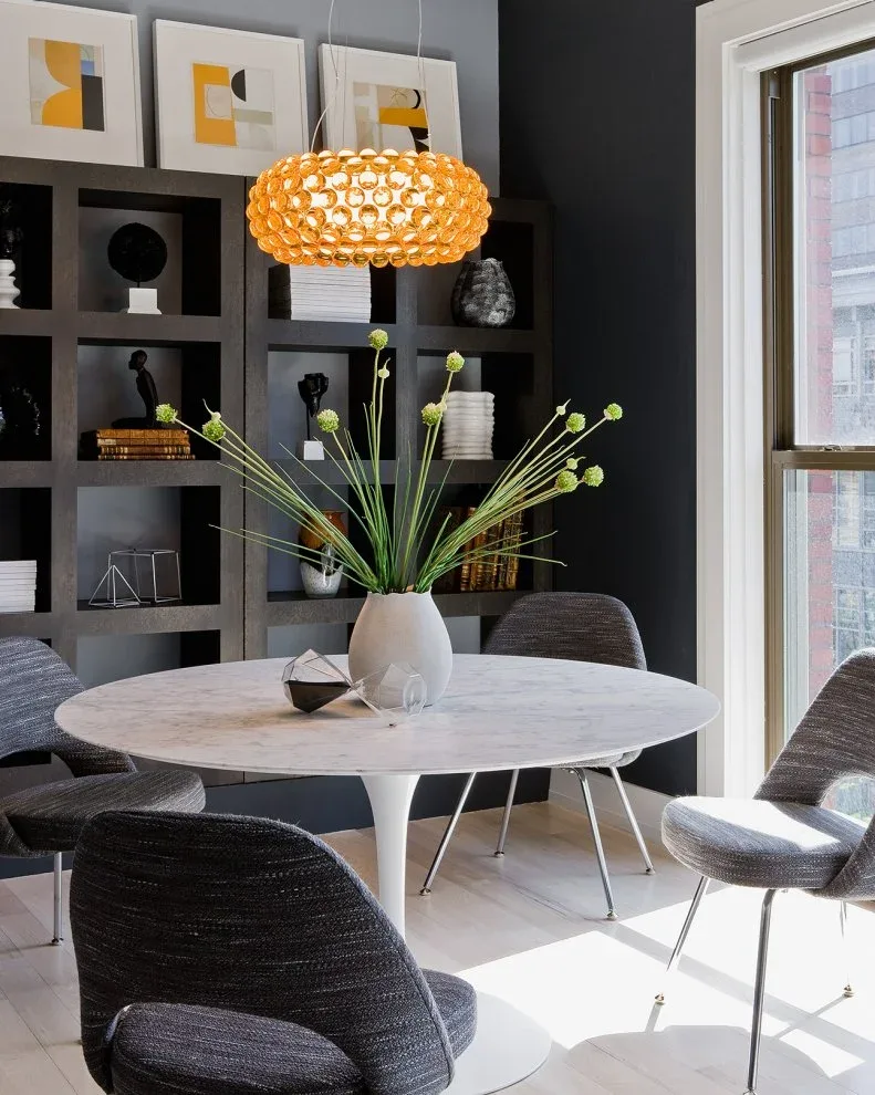 Dark gray upholstered chairs with rounded backs and thin metal legs around a pale round pedestal table, beside black shelving and a broad window.