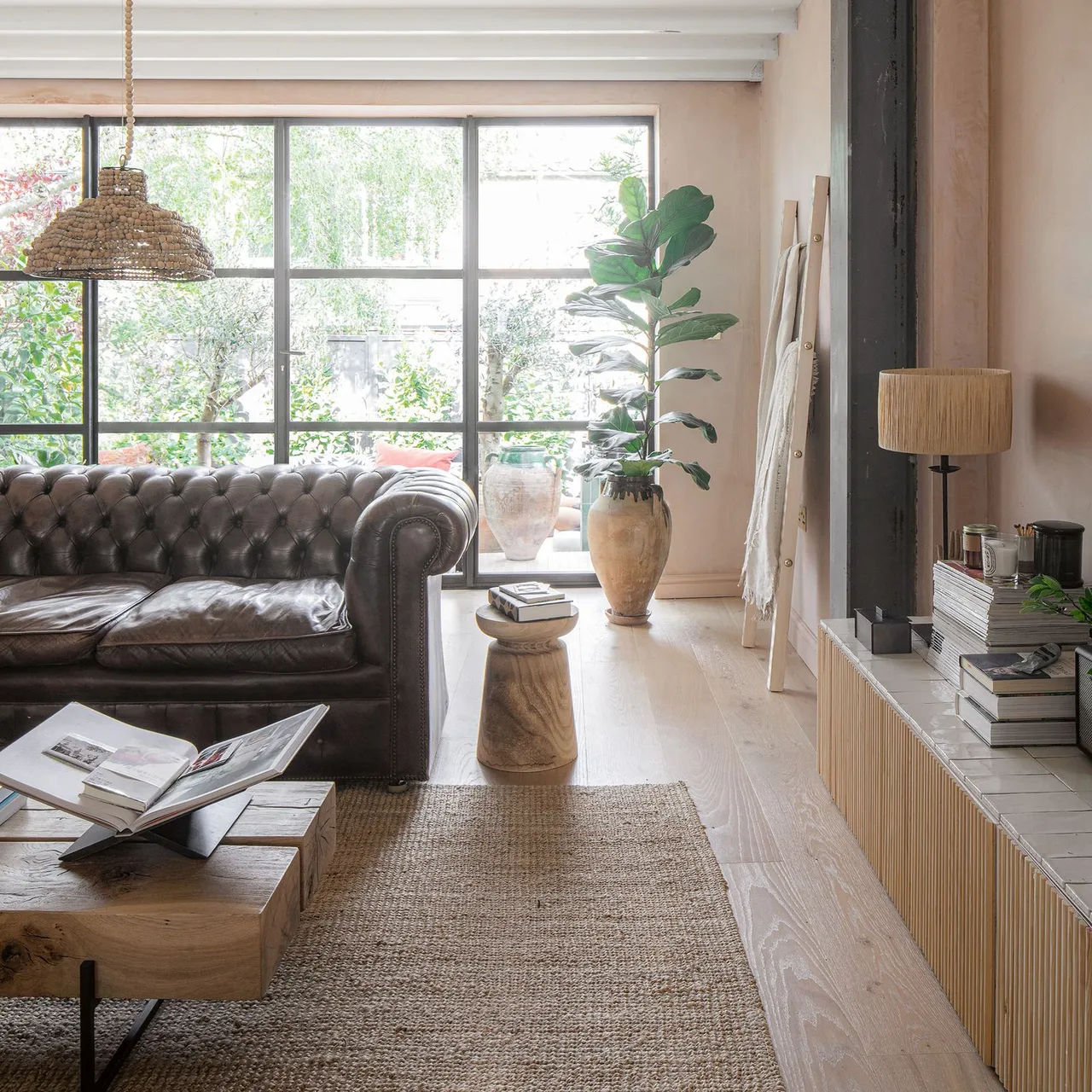 A dark tufted leather sofa sits beside a low wooden coffee table, woven rug, large window and potted plant