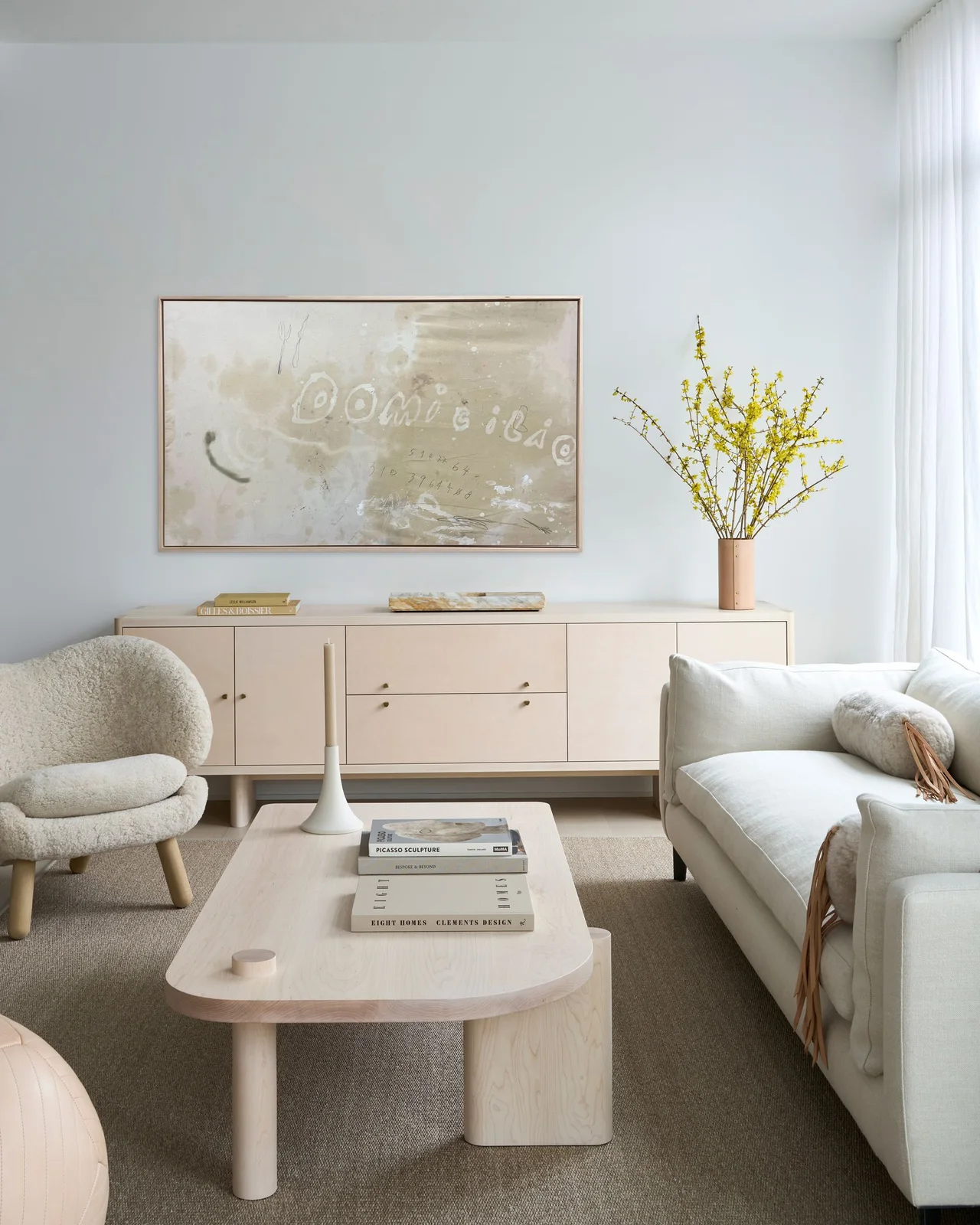 A pale living room has a long sideboard beneath one framed artwork beside a white sofa, rounded chair and pale coffee table