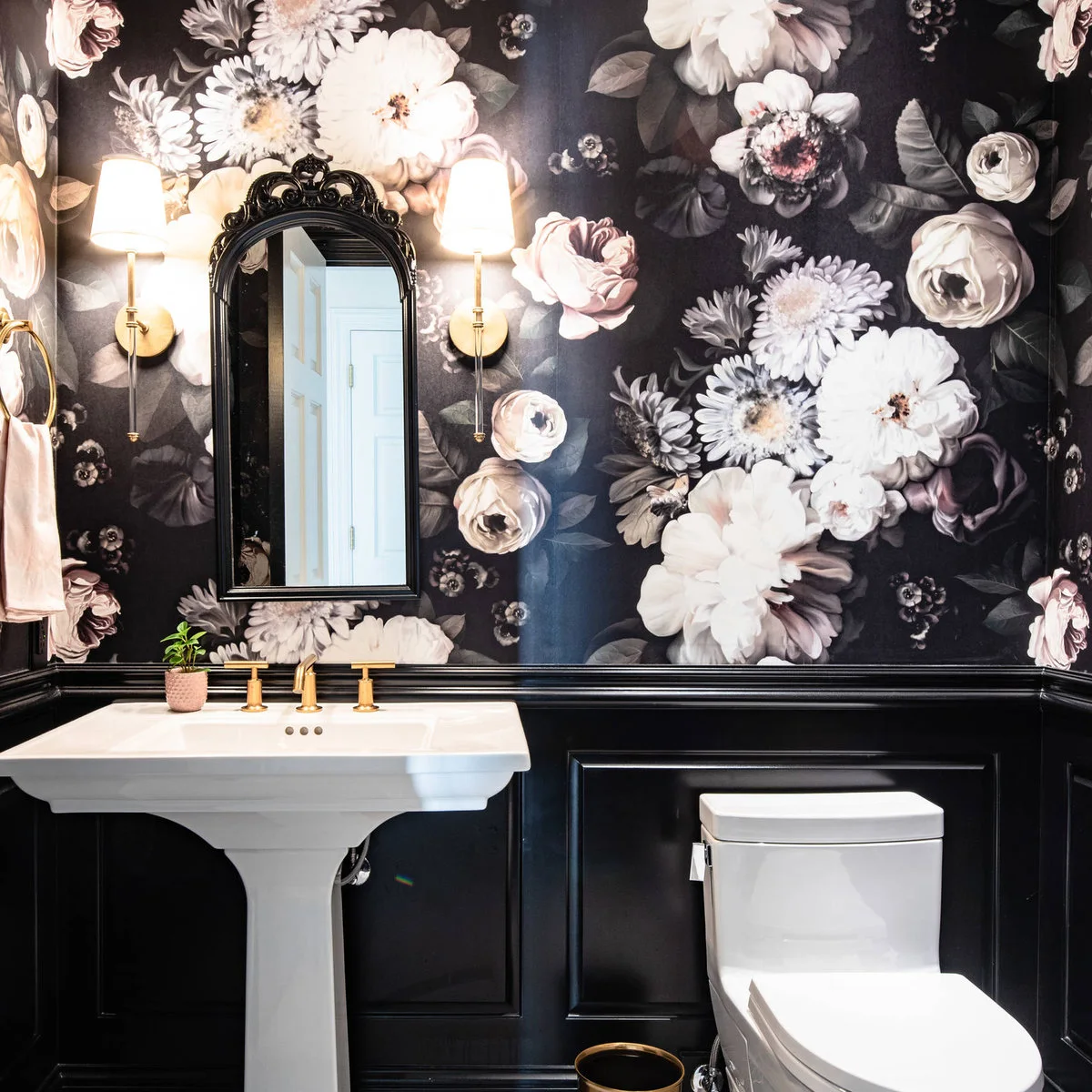 A white pedestal sink sits against black paneling beneath large pale floral wallpaper, with a dark arched mirror and warm metal fixtures.