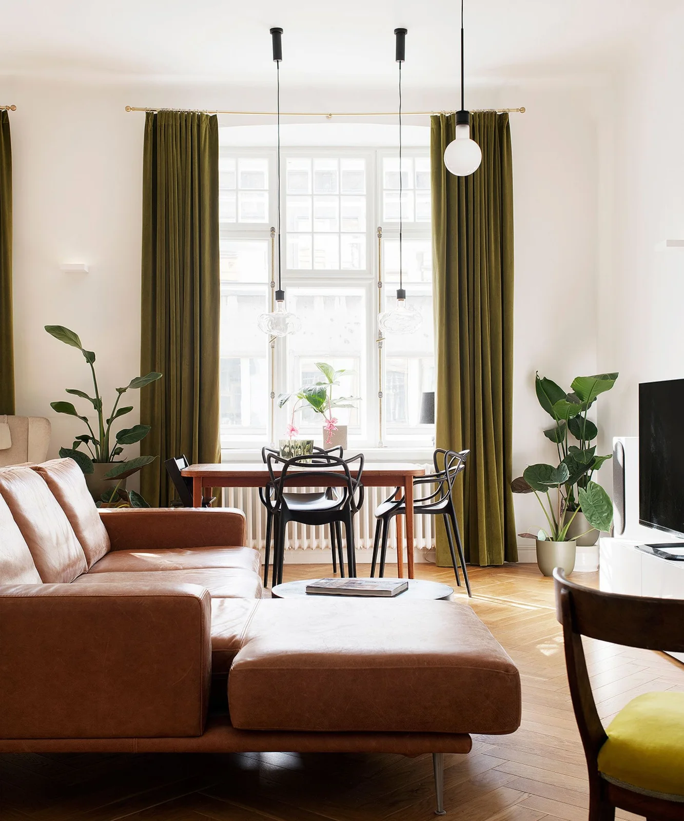 Brown seating and a timber dining table sit beneath olive-green curtains beside leafy plants and pale walls.