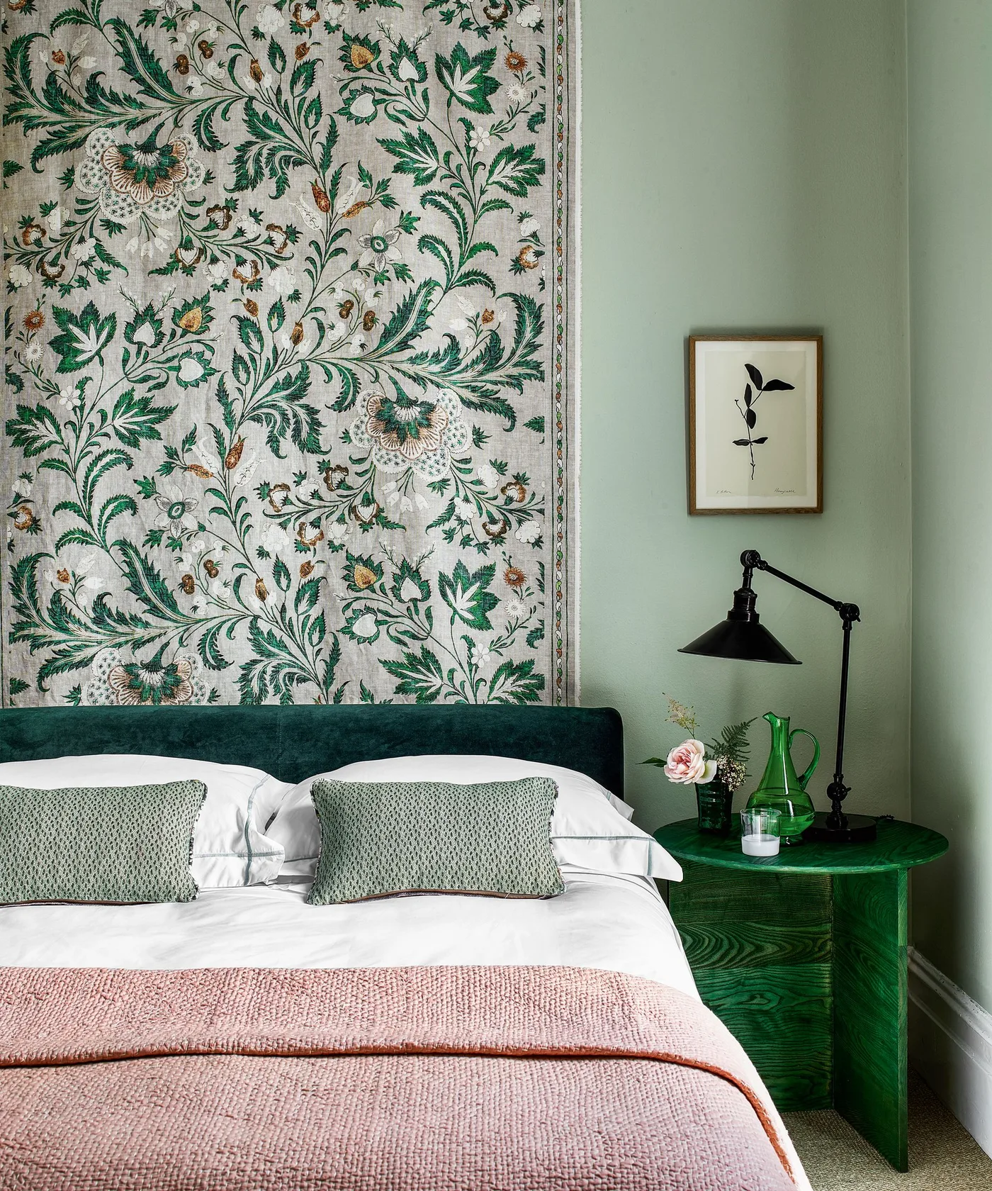 Bedroom with emerald upholstered headboard and round bedside table, pale green wall, botanical wall hanging, white pillows, and pink blanket.