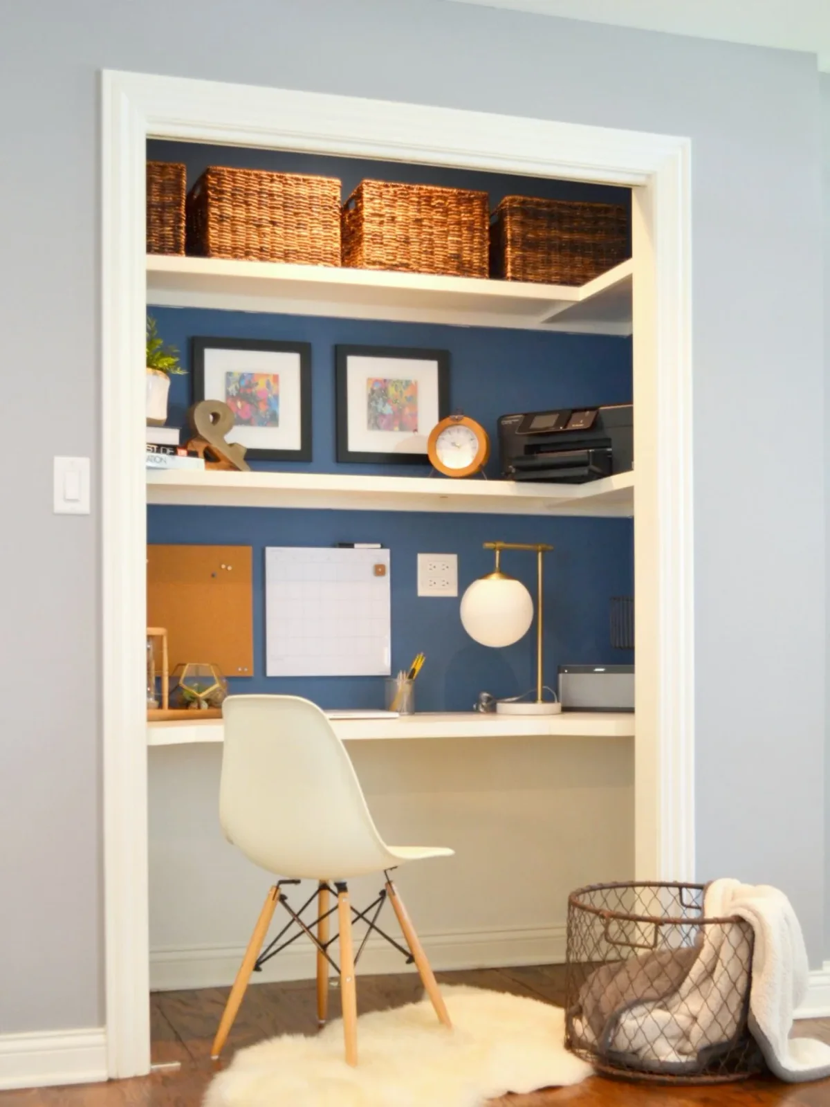 A white desk sits within a blue-backed closet recess beneath shelves and baskets, with a white chair and small globe lamp.