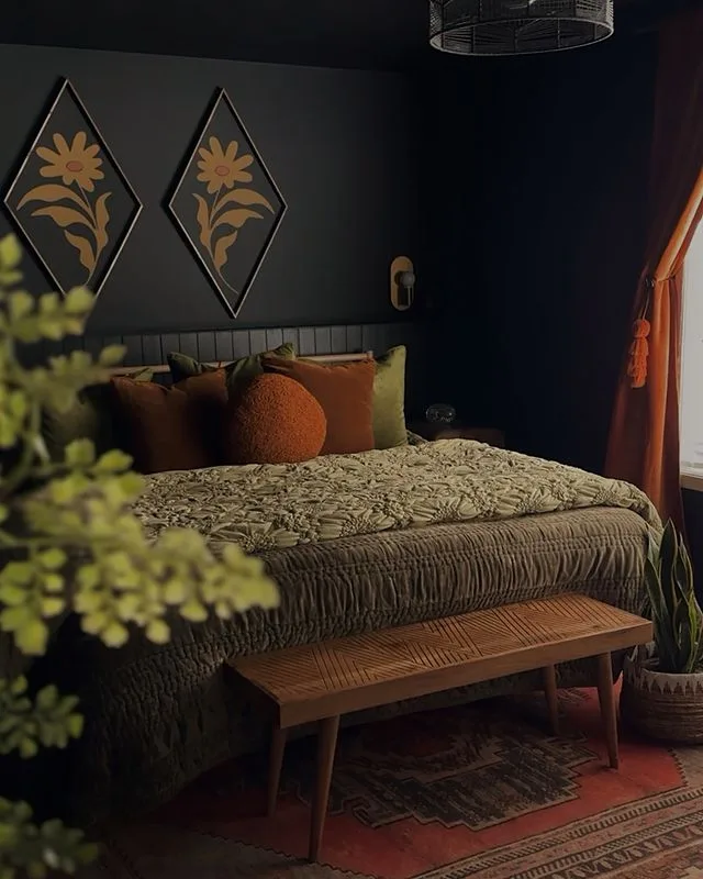 Dark bedroom walls with two diamond-shaped floral pictures, green bedding, warm-colored cushions, wooden bench, and patterned rugs.