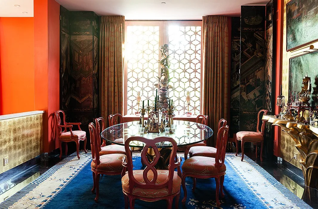 Round glass dining table with red curved chairs, orange walls, dark decorative panels and a blue rug.