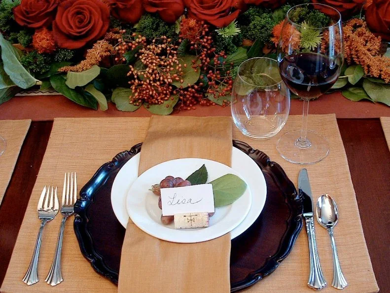 A pale name card stands in a small cork in front of grapes and leaves on a white plate above a dark charger