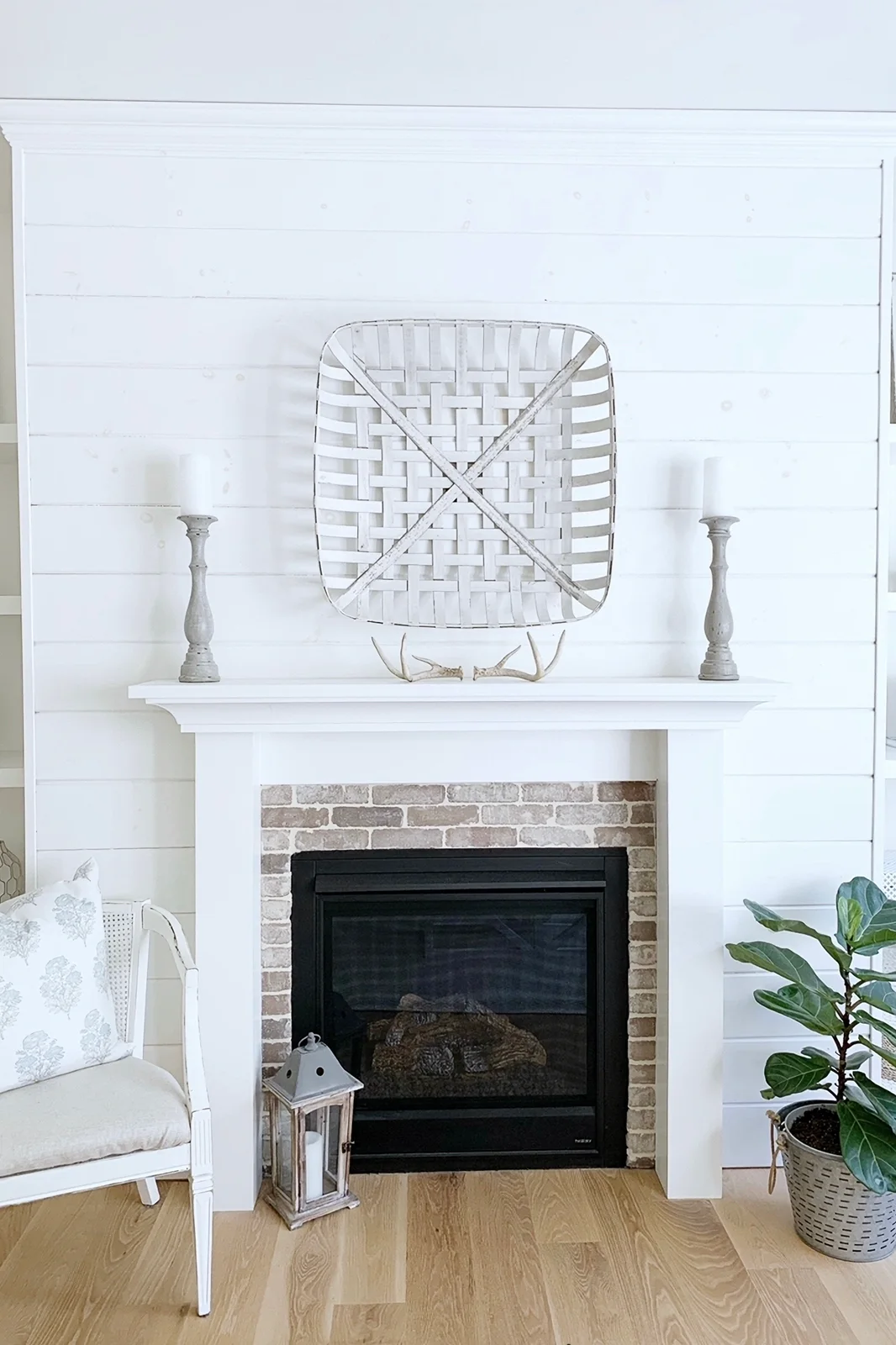 A white shiplap wall surrounds a white mantel and pale brick border, with a dark glazed fireplace, woven wall decoration, paired candlesticks and a small chair.