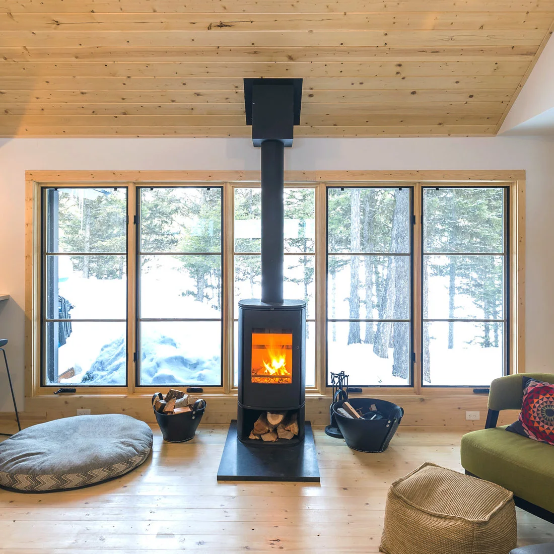 A black cylindrical stove sits on a dark rectangular hearth before wood-trimmed windows, beneath a pale wood ceiling, with green seating and log carriers at the sides.