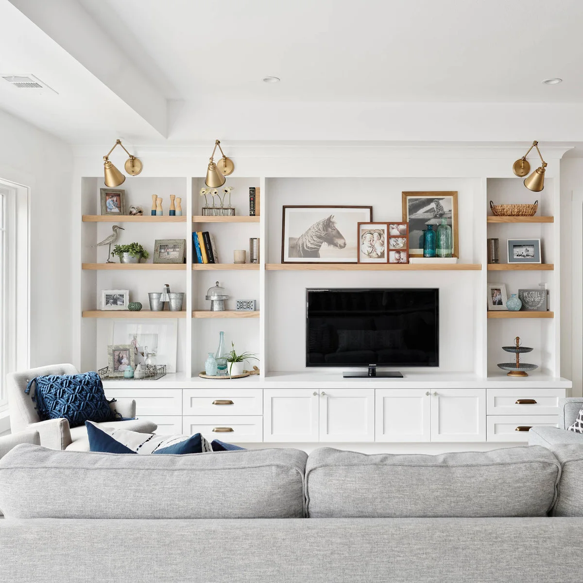 Wide wood shelf with layered framed pictures and blue glass bottles above a television within white built-ins.
