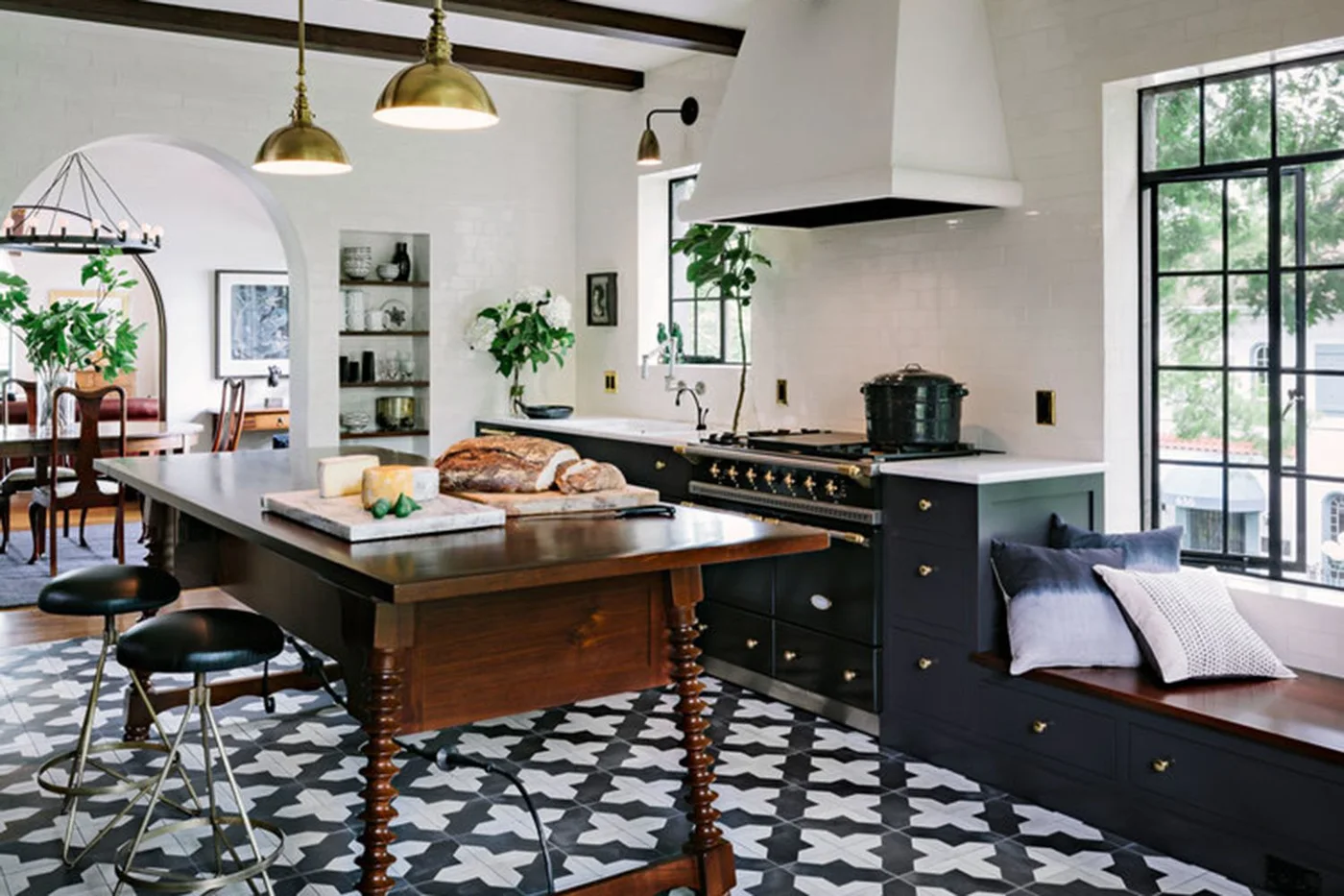 A kitchen has black-and-white patterned flooring beneath a warm wood preparation table beside dark cabinets, white walls, a white hood and black window frames