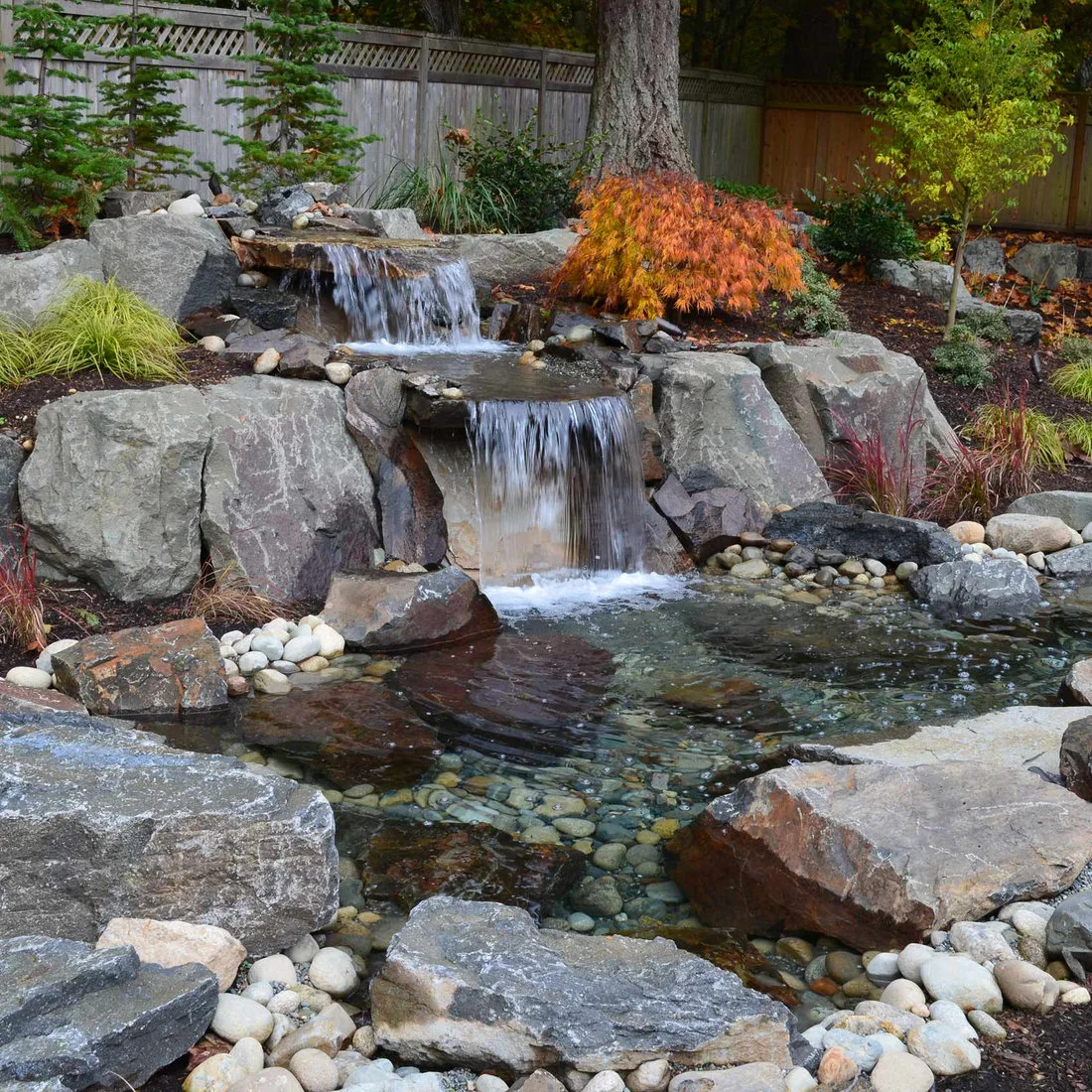 A compact rock waterfall with two broad drops, gray boulders, a pebble-edged pool and an orange-leaved tree behind.