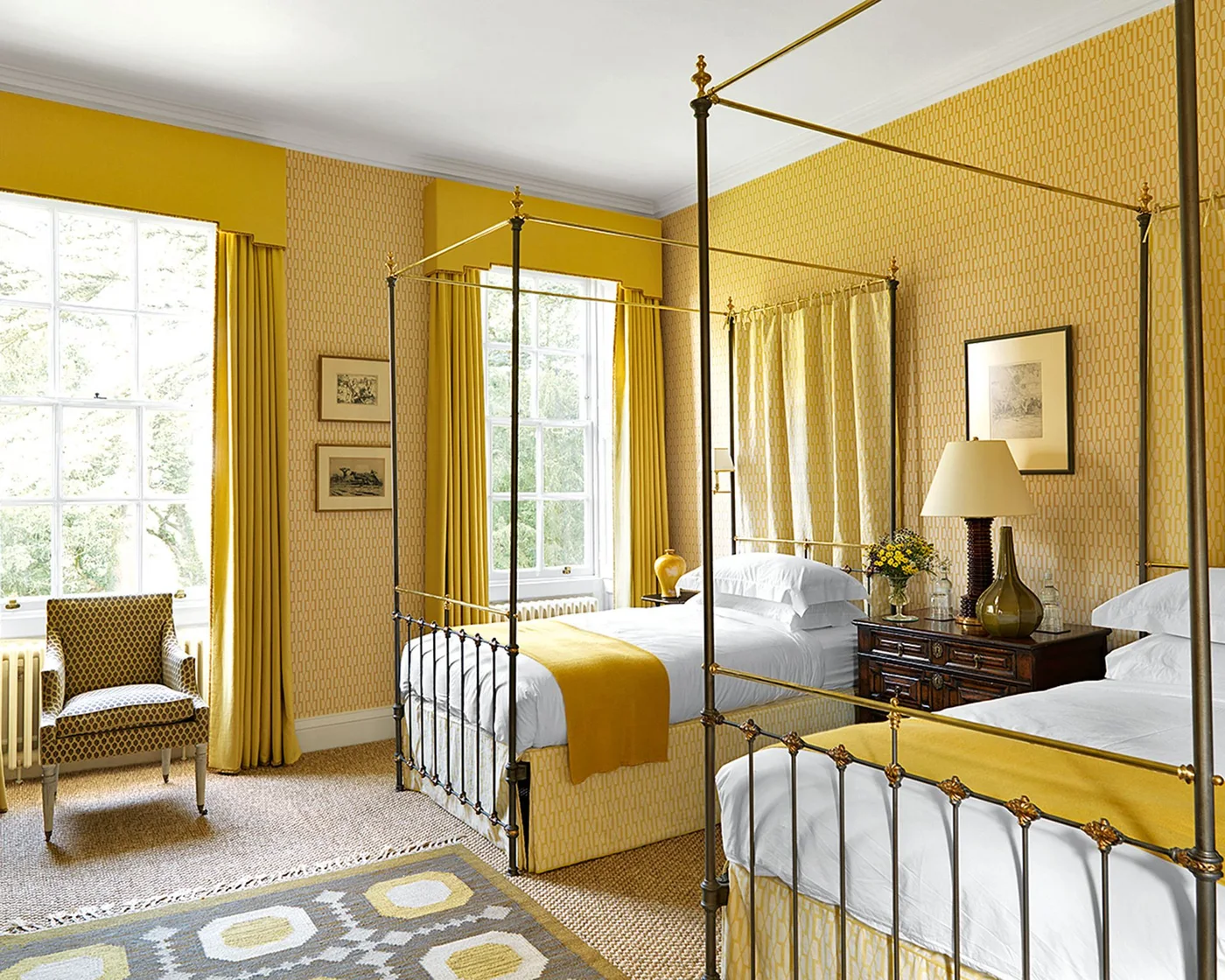 Two metal four-poster beds stand against yellow patterned wall panels with yellow runners, curtains and a shared wood drawer chest.