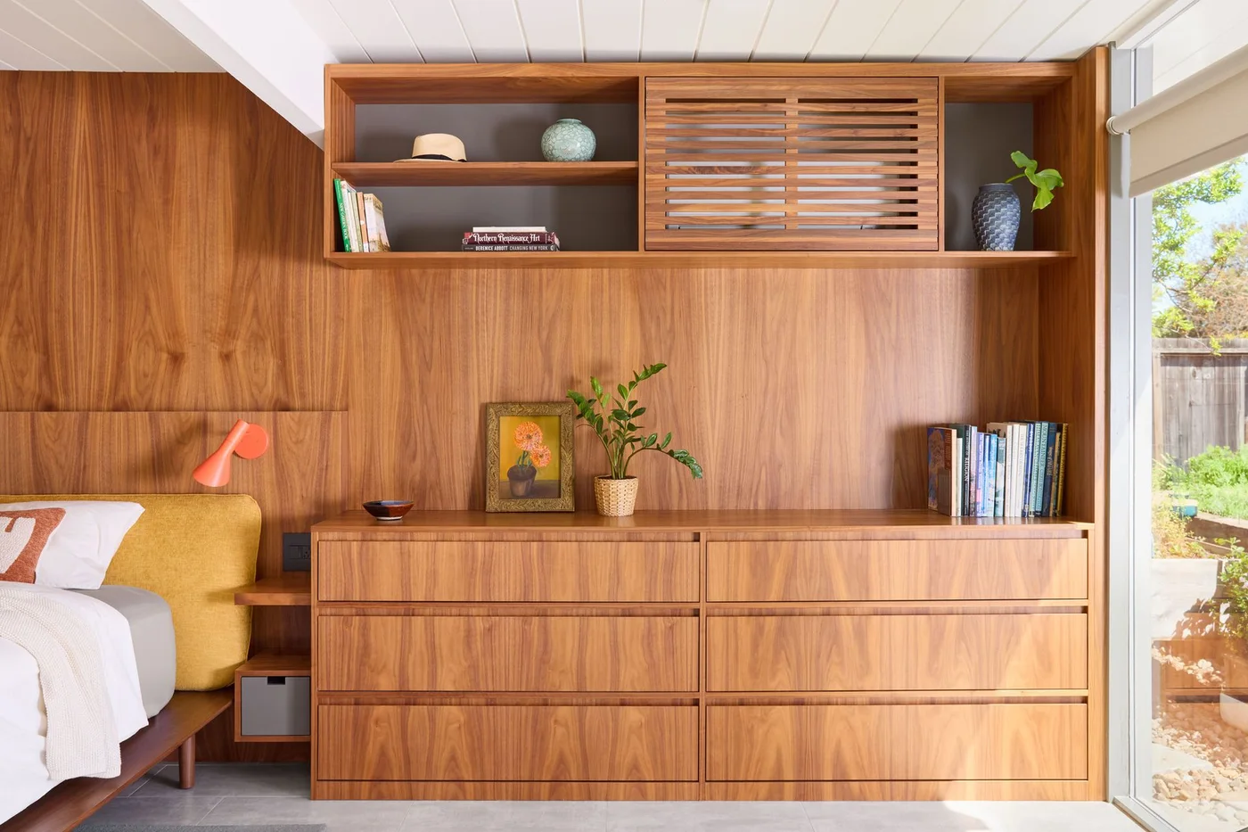 Wood bedroom wall panels continue behind a long drawer cabinet and overhead shelves with a slatted door beside yellow bedding and a glazed opening