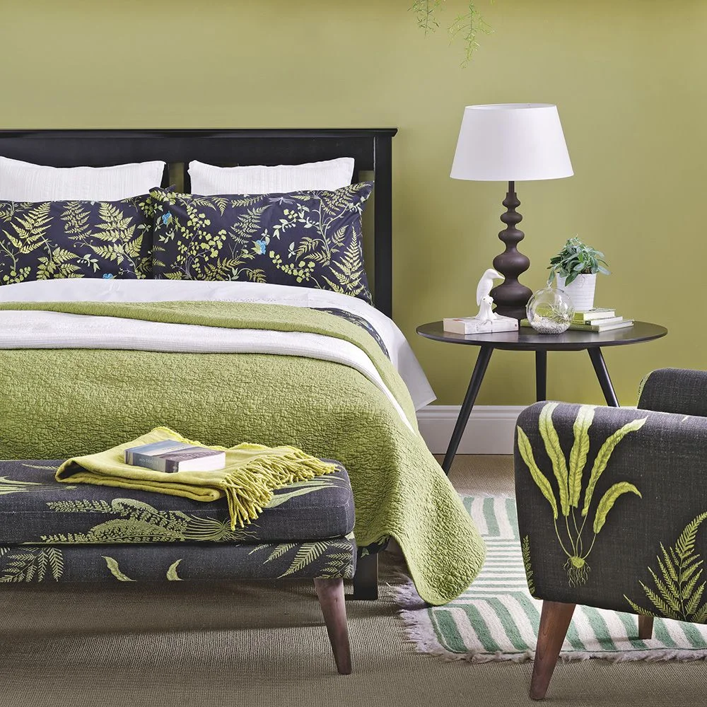 Olive-colored walls surround a dark bed with botanical cushions, a pale green bedspread and dark upholstered seating with green motifs
