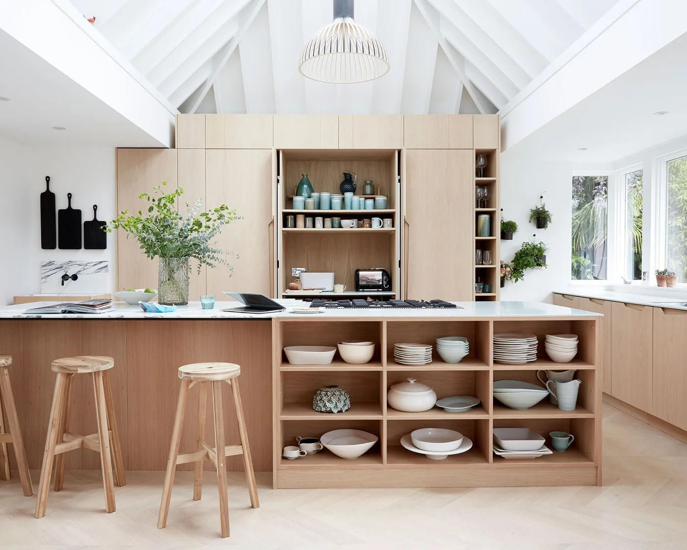 Pale wooden kitchen cabinets have open cubbies holding dishes and a recessed shelf for mugs and appliances